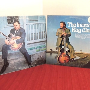 May include: Two vinyl record albums featuring Roy Clark. The left cover shows him sitting on a porch with a guitar, titled Urban, Suburban. The right cover shows him standing by a pond with a guitar, titled The Incredible Roy Clark.