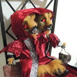May include: A two-headed wolf doll wearing a red velvet hooded cloak with a black spiderweb design. The doll is sitting on a chair with a paisley patterned fabric.