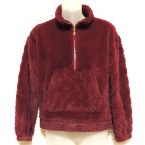 May include: A burgundy faux fur pullover with a quarter-zip closure and a kangaroo pocket. The long-sleeved top has a high collar and a quilted texture on the sleeves and pocket. The zipper is gold-colored.