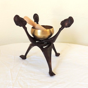 May include: A metallic gold singing bowl with a light-colored wooden striker resting inside, supported by a dark brown carved wooden stand featuring three stylized figures with arms raised to hold the bowl in a central position.