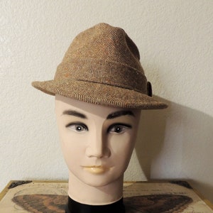 May include: A brown tweed fedora hat with a herringbone pattern. The hat is on a mannequin head.