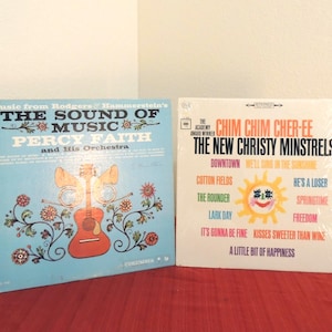 May include: Two vinyl record albums. The left cover is light blue with a red guitar illustration, floral motifs, and text reading Music from Rodgers & Hammerstein's The Sound of Music Percy Faith and His Orchestra. The right cover is white with text Chim Chim Cher-ee The New Christy Minstrels and a yellow sun graphic.