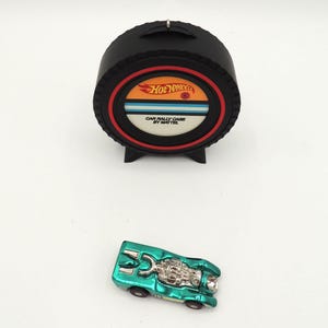 May include: A black Hot Wheels Car Rally Case by Mattel with a red and orange circular design. A teal toy car is next to the case. The case is shaped like a tire.