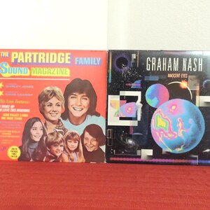 May include: Two vinyl record albums standing side by side. The left album is The Partridge Family Sound Magazine with a red cover featuring a group portrait. The right album is Graham Nash Innocent Eyes with a black cover featuring space graphics.