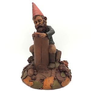 May include: A detailed figurine of a garden gnome, seated on a wooden post. The gnome wears a red pointed hat, a gray jacket, and brown boots. The base features autumn leaves, acorns, and the text "Potter, Ewk 1992".