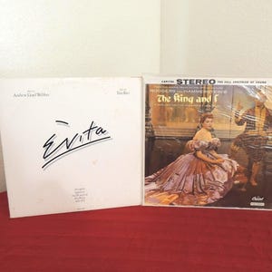 May include: Two vinyl record albums standing side by side. The left album is white with the black text Evita, Andrew Lloyd Webber, and Tim Rice. The right album is The King and I, featuring a color illustration of a person in a large gown.
