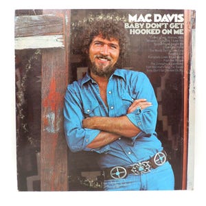 May include: Vintage vinyl record album cover featuring Mac Davis. The album is titled "Baby Don't Get Hooked on Me." The cover shows a man in a denim shirt and jeans, leaning against a wooden structure. The album title and song list are visible.