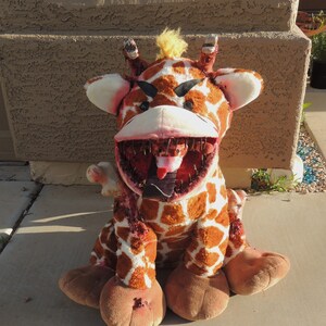 May include: A plush giraffe toy with a distressed, bloody appearance. The giraffe has a large, open mouth with sharp teeth and a long, pink tongue sticking out. The toy is sitting on a concrete surface.