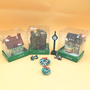 May include: Miniature Christmas village scene featuring three light-up houses with detailed facades and red tile roofs, displayed in clear boxes. Additional elements include a clock tower, benches, and wreaths, all set against a light orange background.