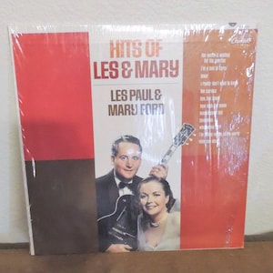 May include: A vinyl record album cover featuring a photo of Les Paul and Mary Ford. The cover is red and brown with the title "Hits of Les & Mary" and "Les Paul & Mary Ford" in white text. The album cover is wrapped in plastic.
