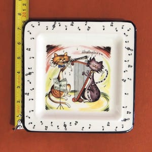May include: A square white ceramic plate featuring a cartoon illustration of an orange tabby cat playing a flute and a grey cat playing a harp. The border is decorated with black musical notes. A yellow measuring tape shows a length of 8 inches.