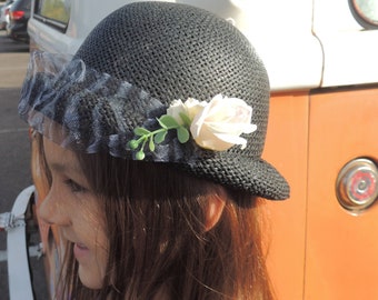 May include: A young person with long brown hair wearing a black and white striped shirt and a black hat with a white flower and a black mesh veil.