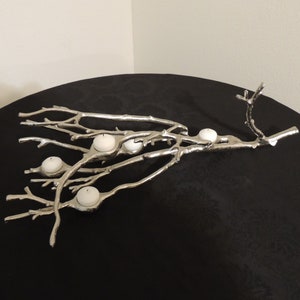 Puede incluir: Portavelas de metal plateado con dise&ntilde;o de rama, con cinco velas blancas. La pieza decorativa est&aacute; colocada sobre una superficie negra, creando un contraste con el metal brillante y las velas.
