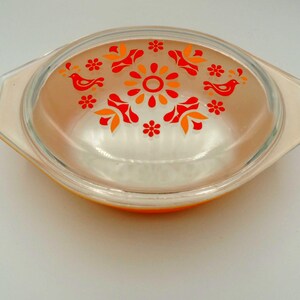 May include: An oval-shaped, vintage Pyrex casserole dish with a clear glass lid. The lid features a floral and bird design in orange and red. The dish has a light orange base and handles.
