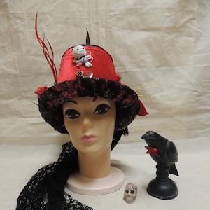 May include: A red and black top hat with a variety of embellishments, including a small doll, a feather, and a skull. The hat is sitting on a mannequin head with a black lace scarf draped around it. A black crow figurine is sitting on a black stand to the right of the hat.