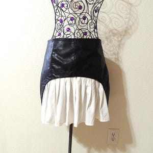 May include: A black faux leather skirt with a white pleated hem is displayed on a decorative wire dress form. The dress form has a black metal stand with a decorative base and is adorned with purple beads.