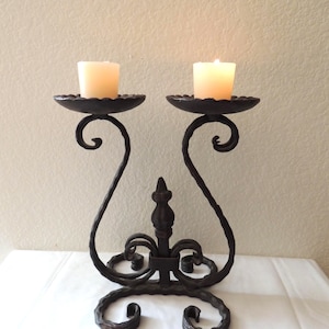 May include: A dark brown, wrought iron double candle holder featuring two circular platforms for votive candles, curved scrollwork supports, and a central decorative finial. Two cream-colored candles are lit and placed on the top platforms.