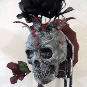 May include: A white skull with red paint and a black rose in its mouth, hanging from a black chain. The skull has a mohawk made of black fabric.