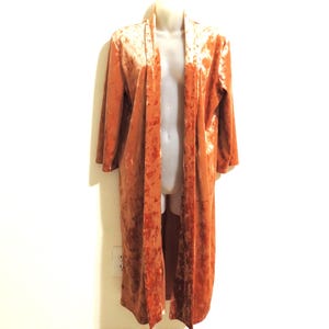 May include: A long, open-front robe in a burnt orange color, made of a plush velvet material. The robe is displayed on a mannequin, showcasing its flowing design and long sleeves. The garment appears to be floor-length.