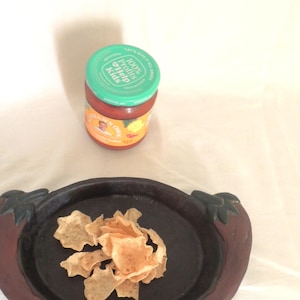 May include: A brown ceramic chip and dip dish with carved handles in the shape of leaves. The dish is filled with a small pile of tortilla chips. A jar of salsa with a green lid is in the background.