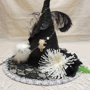 May include: A black witch hat with a pointed crown, adorned with white beads, a white feather boa, a white flower, and a black feather. The hat is trimmed with black lace and has a white skull charm.