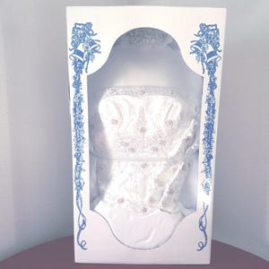 May include: A white wedding dress with lace and beading details is displayed in a white box with blue floral accents and the words "Wedding Bells".