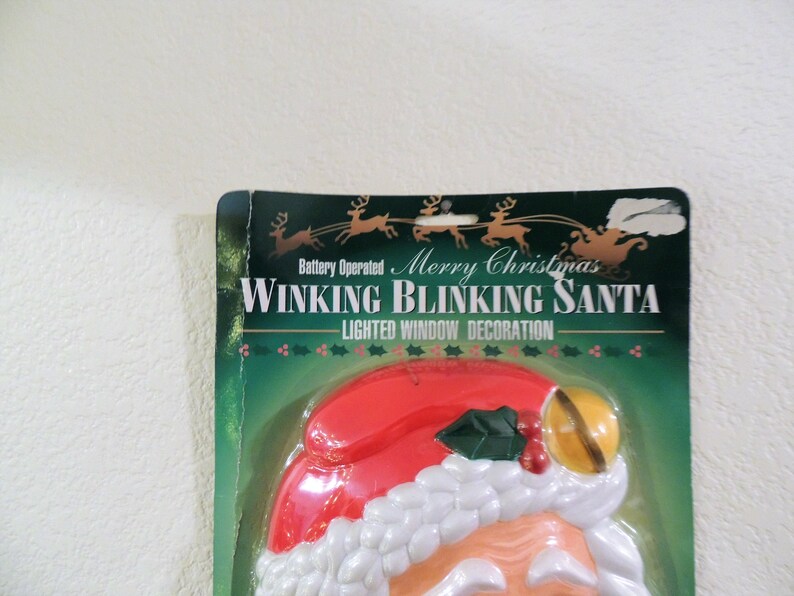 May include: A plastic, battery-operated, lighted window decoration of Santa Claus with a red hat, white beard, and a bell. The packaging says "Merry Christmas Winking Blinking Santa Lighted Window Decoration".