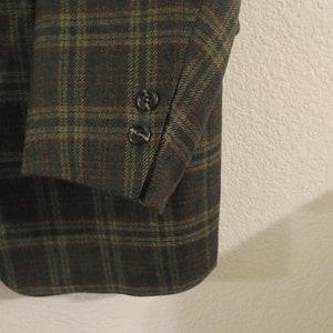 May include: A close-up of the sleeve of a plaid blazer. The blazer is made of a dark green and brown fabric with a subtle texture. The sleeve has two buttons at the cuff.