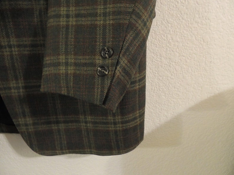 May include: A close-up of the sleeve of a plaid blazer. The blazer is made of a dark green and brown fabric with a subtle texture. The sleeve has two buttons at the cuff.