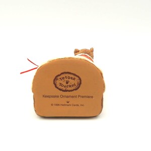 Może przedstawiać: Brązowa figurka Hallmark Keepsake Ornament Premiere przedstawiająca małego psa na brązowej podstawie. Podstawa zawiera napis "Tender Touches" i "1996 Hallmark Cards, Inc." Dołączona jest czerwona wstążka.