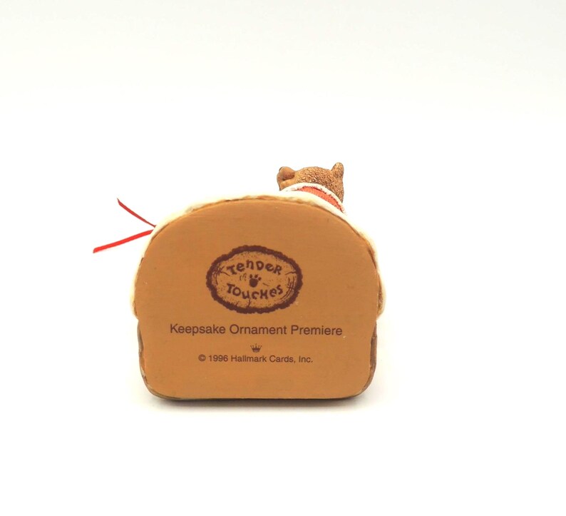 Może przedstawiać: Brązowa figurka Hallmark Keepsake Ornament Premiere przedstawiająca małego psa na brązowej podstawie. Podstawa zawiera napis "Tender Touches" i "1996 Hallmark Cards, Inc." Dołączona jest czerwona wstążka.