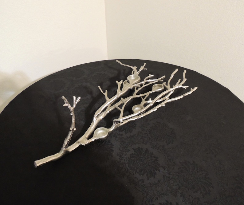 Puede incluir: Candelabro plateado en forma de rama con m&uacute;ltiples huecos para velas. La pieza decorativa es de metal y tiene un dise&ntilde;o naturalista. El candelabro est&aacute; colocado sobre una superficie negra.