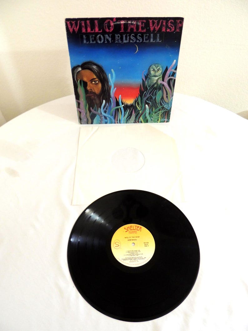 May include: A vintage vinyl record album titled "Will O' The Wisp" by Leon Russell. The album cover features a painted illustration with a portrait, an owl, and the text "Will O' The Wisp Leon Russell". The record is black with a yellow label.
