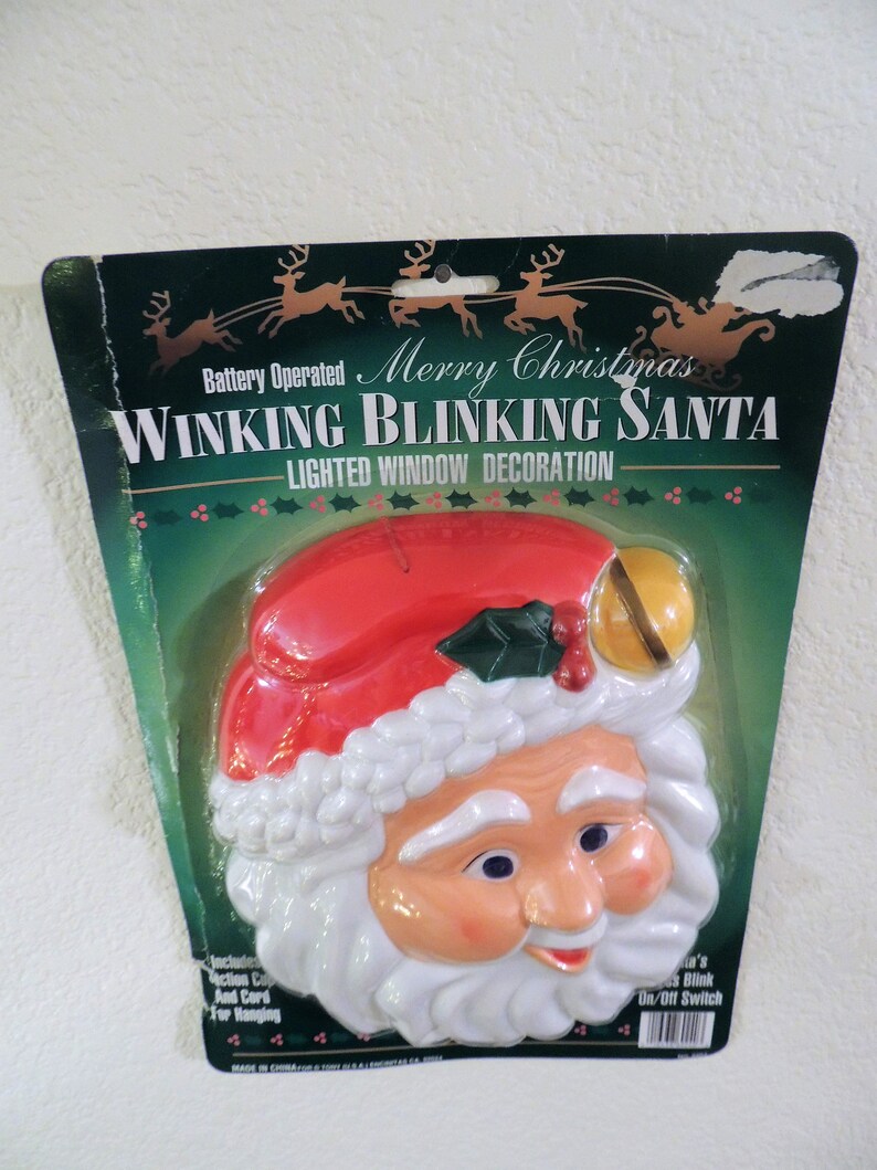 May include: A plastic, white Santa Claus face with a red hat and a green holly sprig. The Santa Claus face is winking and blinking with battery-operated lights. The packaging says "Merry Christmas Winking Blinking Santa - Lighted Window Decoration".