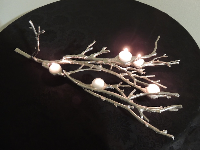 Puede incluir: Un candelabro plateado en forma de rama con seis velas blancas. Las velas est&aacute;n encendidas, emitiendo un brillo c&aacute;lido. El soporte es de metal y se encuentra sobre una superficie negra. El dise&ntilde;o se asemeja a una rama de &aacute;rbol natural.