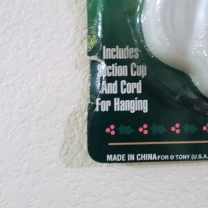 May include: A white ceramic Santa Claus ornament with a green background. The ornament includes a suction cup and cord for hanging. The text on the background reads "Includes Suction Cup And Cord For Hanging" and "MADE IN CHINA FOR @TONY (U.S.A.)".