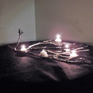 Puede incluir: Un centro de mesa decorativo con un dise&ntilde;o de ramas plateadas y varias velas blancas encendidas. Las velas est&aacute;n colocadas sobre las ramas, creando un brillo c&aacute;lido. El arreglo se encuentra sobre una superficie oscura y texturizada.