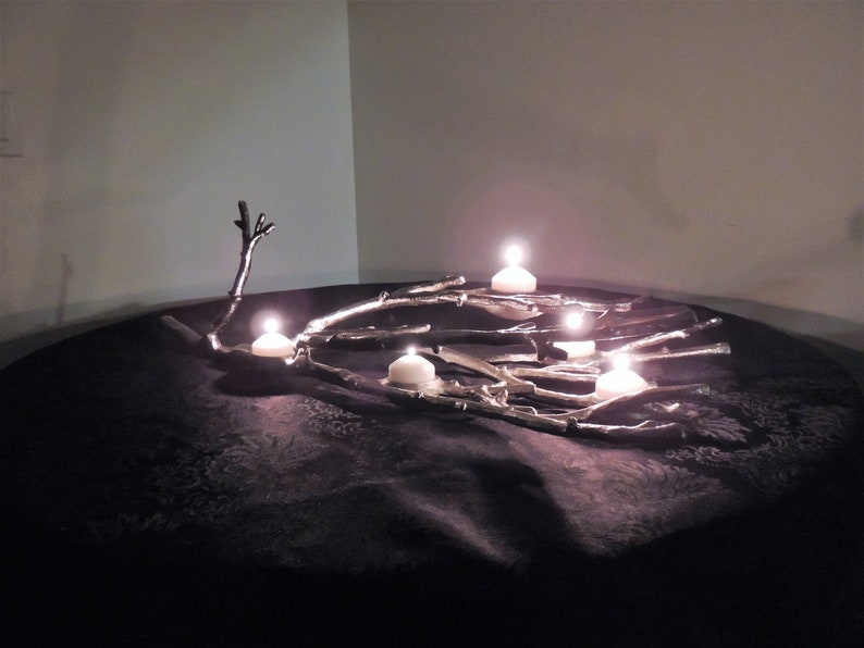 Puede incluir: Un centro de mesa decorativo con un dise&ntilde;o de ramas plateadas y varias velas blancas encendidas. Las velas est&aacute;n colocadas sobre las ramas, creando un brillo c&aacute;lido. El arreglo se encuentra sobre una superficie oscura y texturizada.
