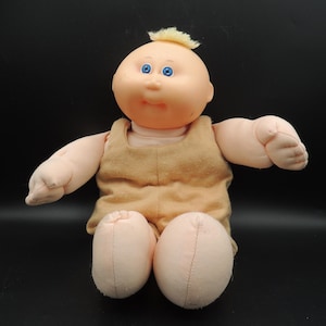 May include: A vintage doll with a peach-colored face, blue eyes, and a small tuft of yellow hair. The doll is wearing a tan outfit and has soft, rounded limbs. The doll is sitting with its arms outstretched.