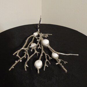 Puede incluir: Candelabro de metal plateado con dise&ntilde;o de ramas, sosteniendo varias velas blancas. El candelabro est&aacute; colocado sobre una superficie negra, creando un contraste con los elementos plateados y blancos. El dise&ntilde;o es intrincado y decorativo.