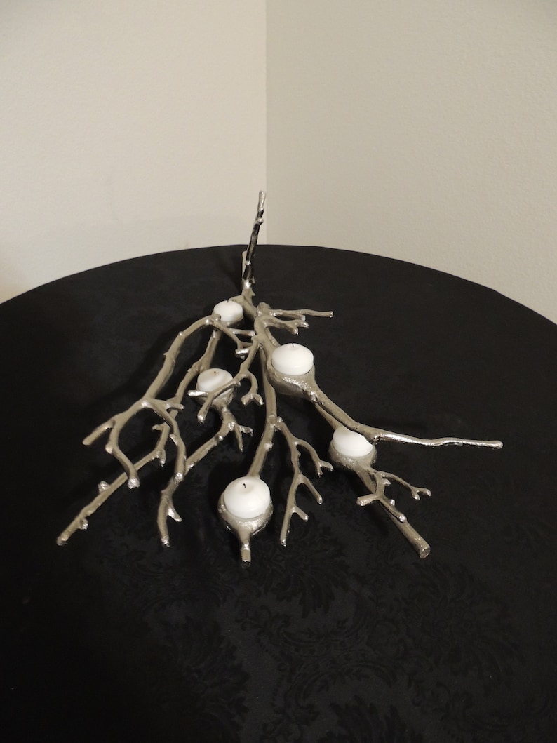 Puede incluir: Candelabro de metal plateado con dise&ntilde;o de ramas, sosteniendo varias velas blancas. El candelabro est&aacute; colocado sobre una superficie negra, creando un contraste con los elementos plateados y blancos. El dise&ntilde;o es intrincado y decorativo.