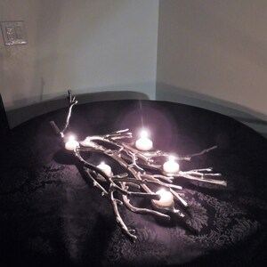 Puede incluir: Un candelabro plateado en forma de rama con cinco velas blancas encendidas. El candelabro est&aacute; sobre una superficie oscura estampada. Las velas son redondas y emiten un brillo c&aacute;lido.