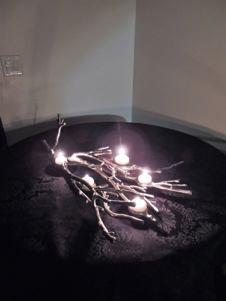 Puede incluir: Un candelabro plateado en forma de rama con cinco velas blancas encendidas. El candelabro est&aacute; sobre una superficie oscura estampada. Las velas son redondas y emiten un brillo c&aacute;lido.