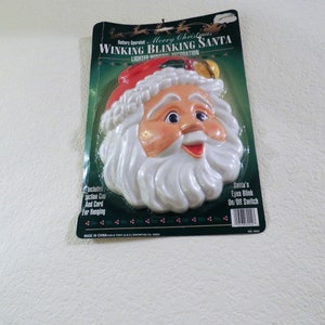 May include: A battery-operated, lighted Christmas decoration featuring a winking Santa Claus head. The Santa has a red hat, white beard, and a jolly expression. The packaging includes the text "Merry Christmas Winking Blinking Santa" and "Santa's Eyes Blink On/Off Switch".