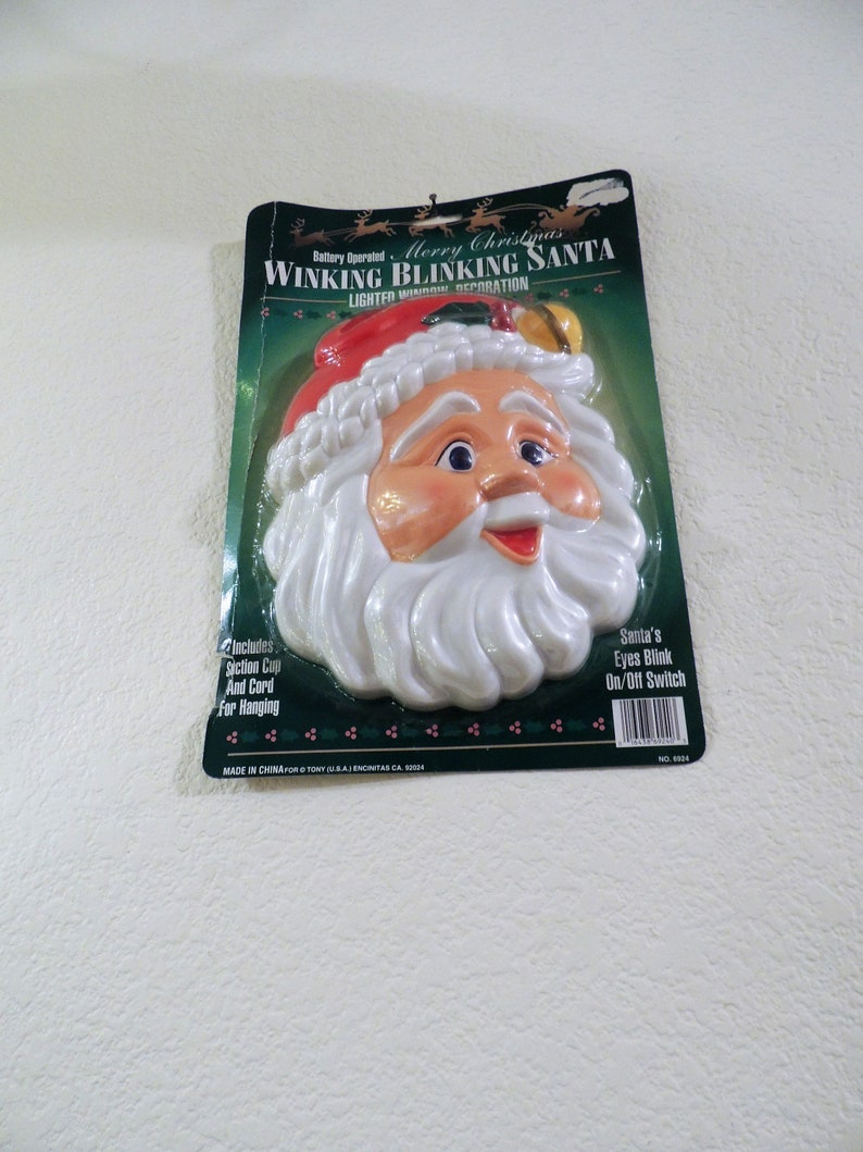May include: A battery-operated, lighted Christmas decoration featuring a winking Santa Claus head. The Santa has a red hat, white beard, and a jolly expression. The packaging includes the text "Merry Christmas Winking Blinking Santa" and "Santa's Eyes Blink On/Off Switch".