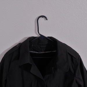 May include: A black trench coat hanging on a black hanger against a white wall.