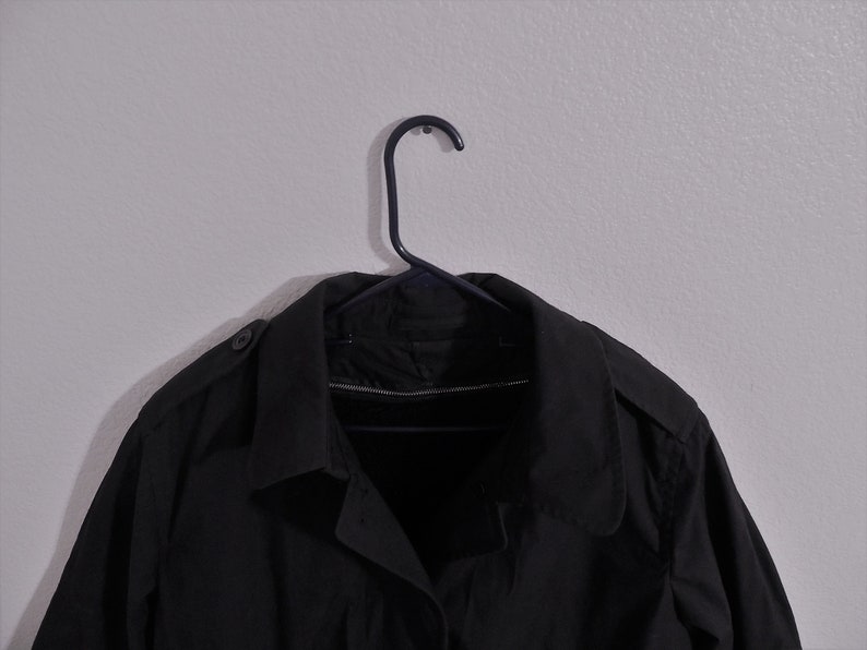 May include: A black trench coat hanging on a black hanger against a white wall.