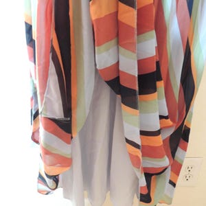 May include: A flowing dress with a colorful striped pattern in shades of orange, black, white, and light green. The dress has a light purple lining and is hanging near a wall with electrical outlets.