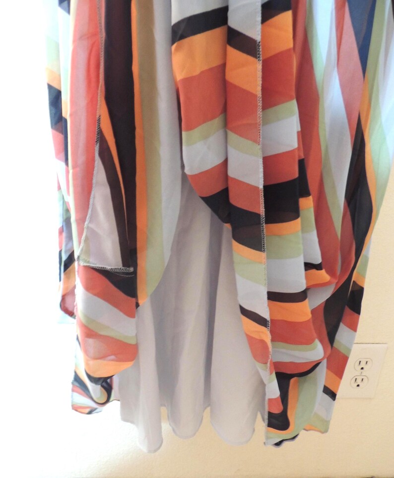 May include: A flowing dress with a colorful striped pattern in shades of orange, black, white, and light green. The dress has a light purple lining and is hanging near a wall with electrical outlets.