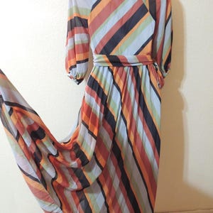 May include: A flowing, vintage maxi dress featuring a vibrant striped pattern in shades of orange, red, black, blue, and green. The dress has long sleeves, a cinched waist, and a pleated skirt, creating a flattering silhouette. The fabric appears lightweight and semi-sheer.